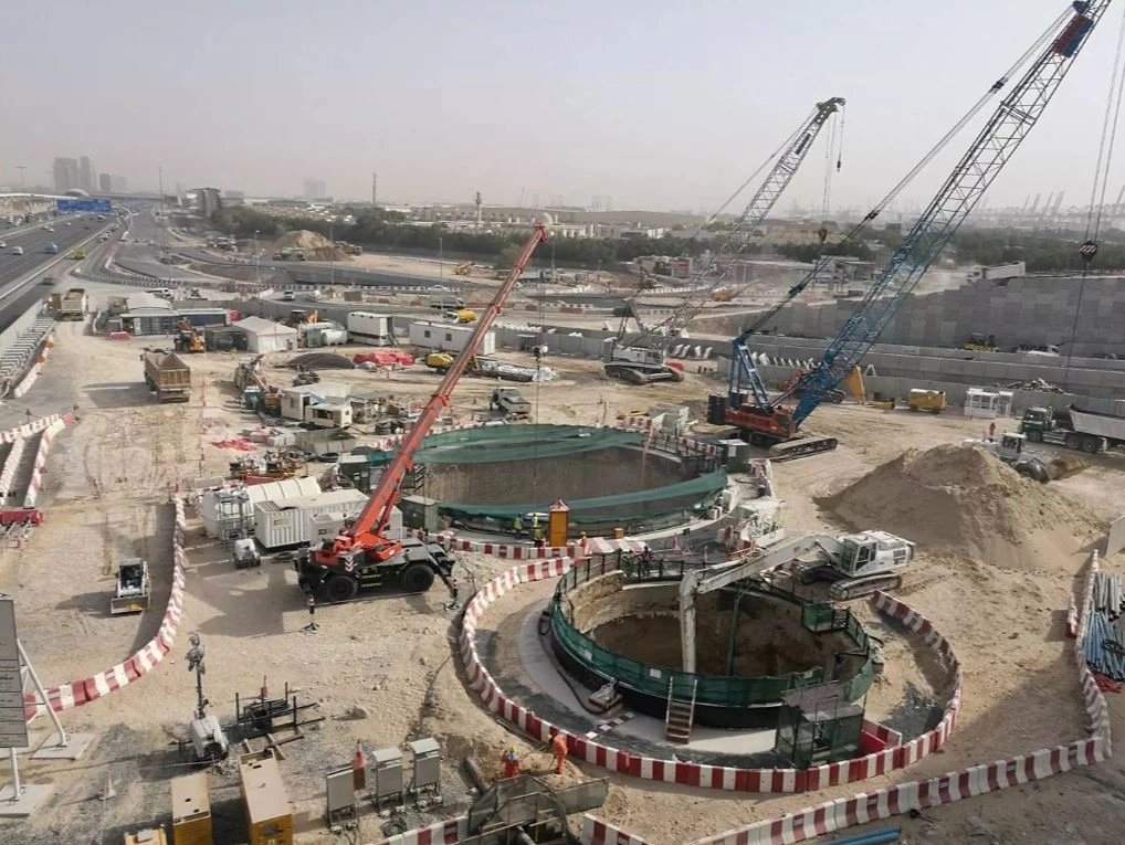 Major transport infrastructure interchange construction showing deep circular shaft excavations, precast retaining wall, cranes, and adjacent live highway